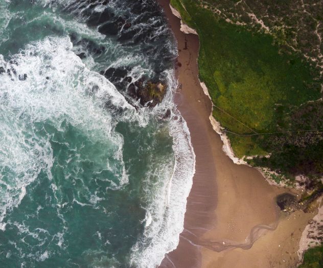 An aerial view of a rocky coastline with waves crashing against the shore, a sandy beach, and green grassy areas surrounding it.