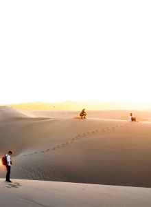 Three people in a vast desert landscape, marked by footprints on the sand dunes under a bright sunrise or sunset, are in the image.