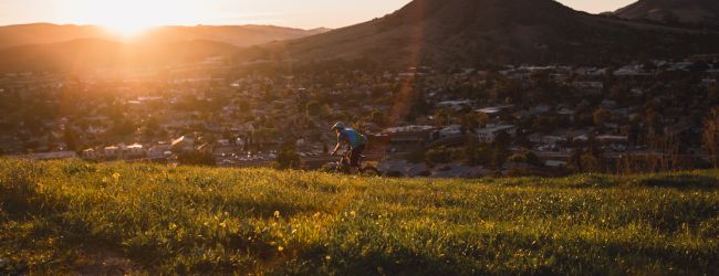 A person is riding a bicycle on a grassy hill with a city and mountains in the background during sunset.