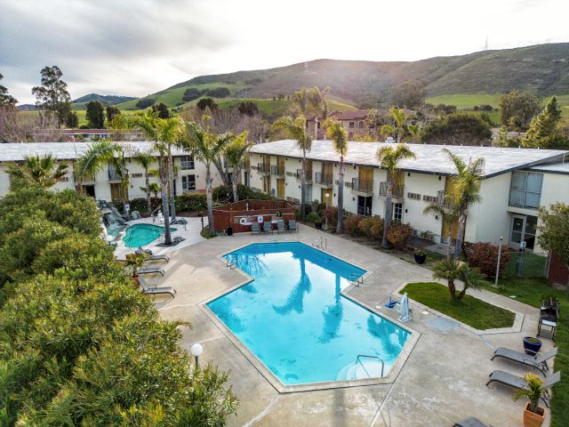 Aerial view of a hotel pool area with a large pool, lounge chairs, and palm trees, surrounded by white buildings and a mountain view.