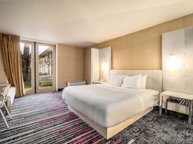 A wide-angle view of a hotel room with a large bed, a door leading outside, and a window.