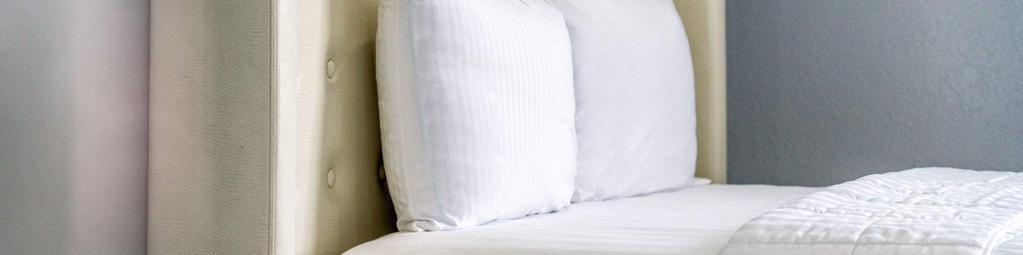 The image shows a neatly made bed with a padded headboard, white pillows, and white bedding against a blue and gray wall.