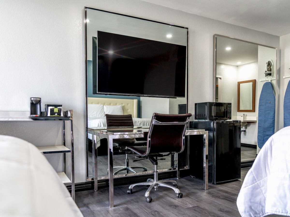 A modern hotel room with a flat-screen TV, desk, ergonomic chair, mini fridge, wall mirror, and ironing board, featuring a clean, minimalist design.