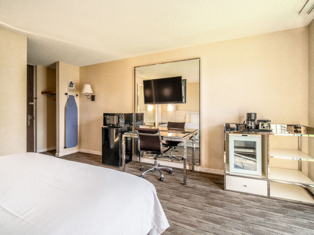 A hotel room with a bed, desk, chair, TV, mini-fridge, microwave, coffee maker, and an ironing board in the corner.