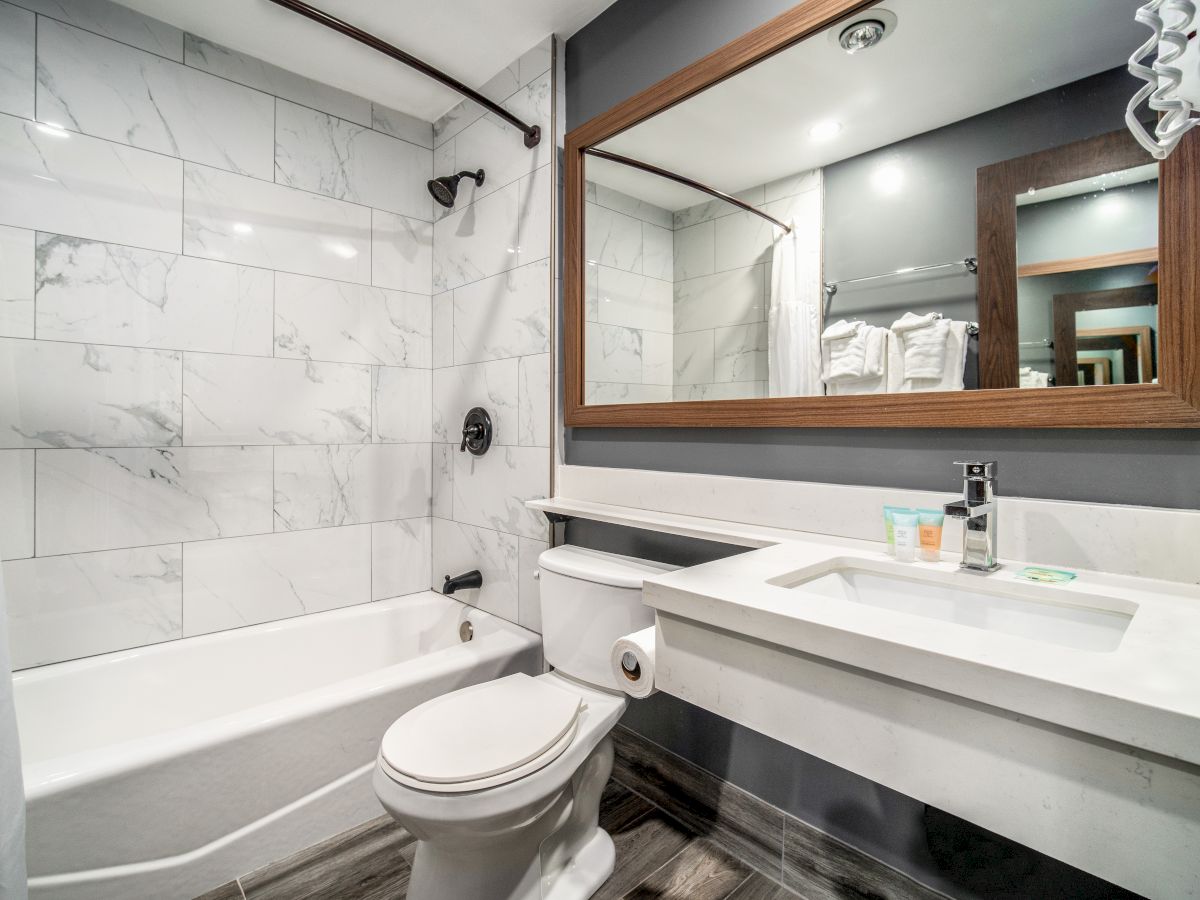 A modern bathroom with a bathtub, toilet, large mirror, and sink, featuring a marble design, wooden accents, and neatly arranged toiletries.