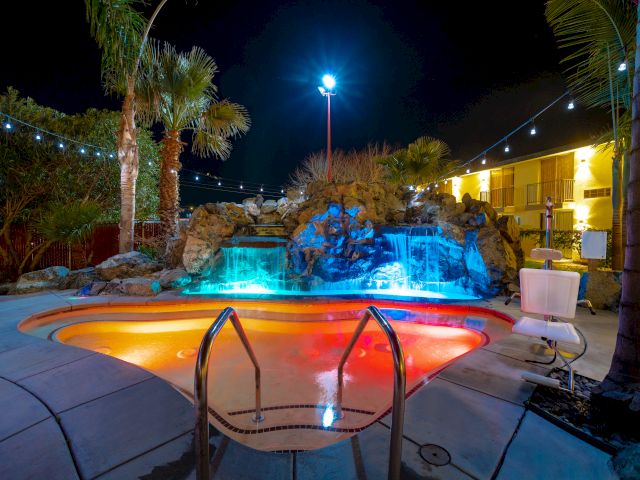 A nighttime view of a hotel pool with colorful lighting, a waterfall, and palm trees.