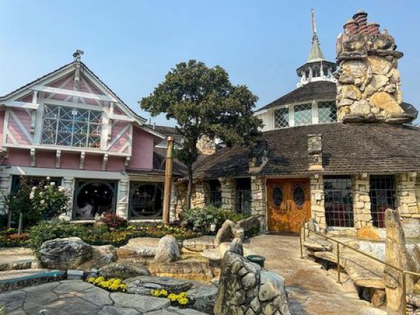 A quaint, fairy-tale-like building with stone and wood architecture, featuring a garden and a pathway leading to the entrance.