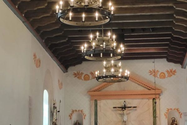 The image shows the interior of a church with lit chandeliers, religious statues, and a crucifix at the altar, beneath a wooden beamed ceiling.