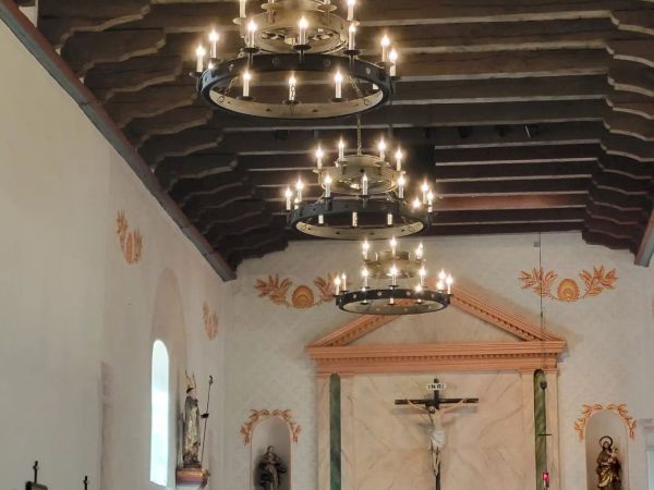 The image shows the interior of a church with lit chandeliers, religious statues, and a crucifix at the altar, beneath a wooden beamed ceiling.