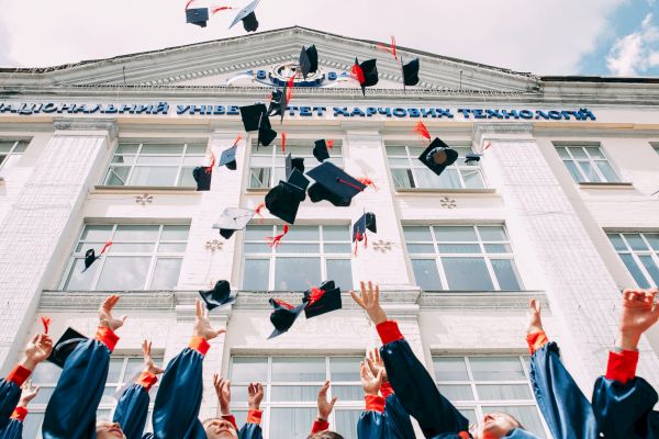 A group of graduates in blue gowns and caps is celebrating by throwing their caps into the air in front of a building.