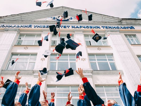 A group of graduates in blue gowns and caps is celebrating by throwing their caps into the air in front of a building.
