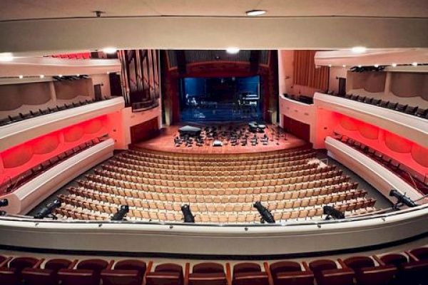 The image shows the interior of a large theater or concert hall with rows of empty seats and a stage at the front.