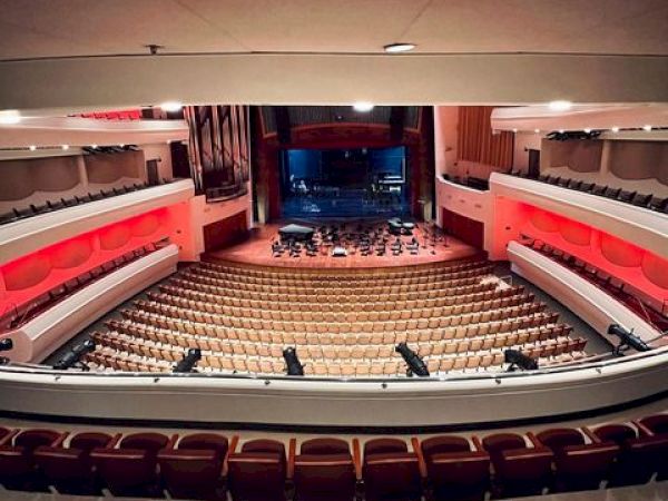 The image shows the interior of a large theater or concert hall with rows of empty seats and a stage at the front.