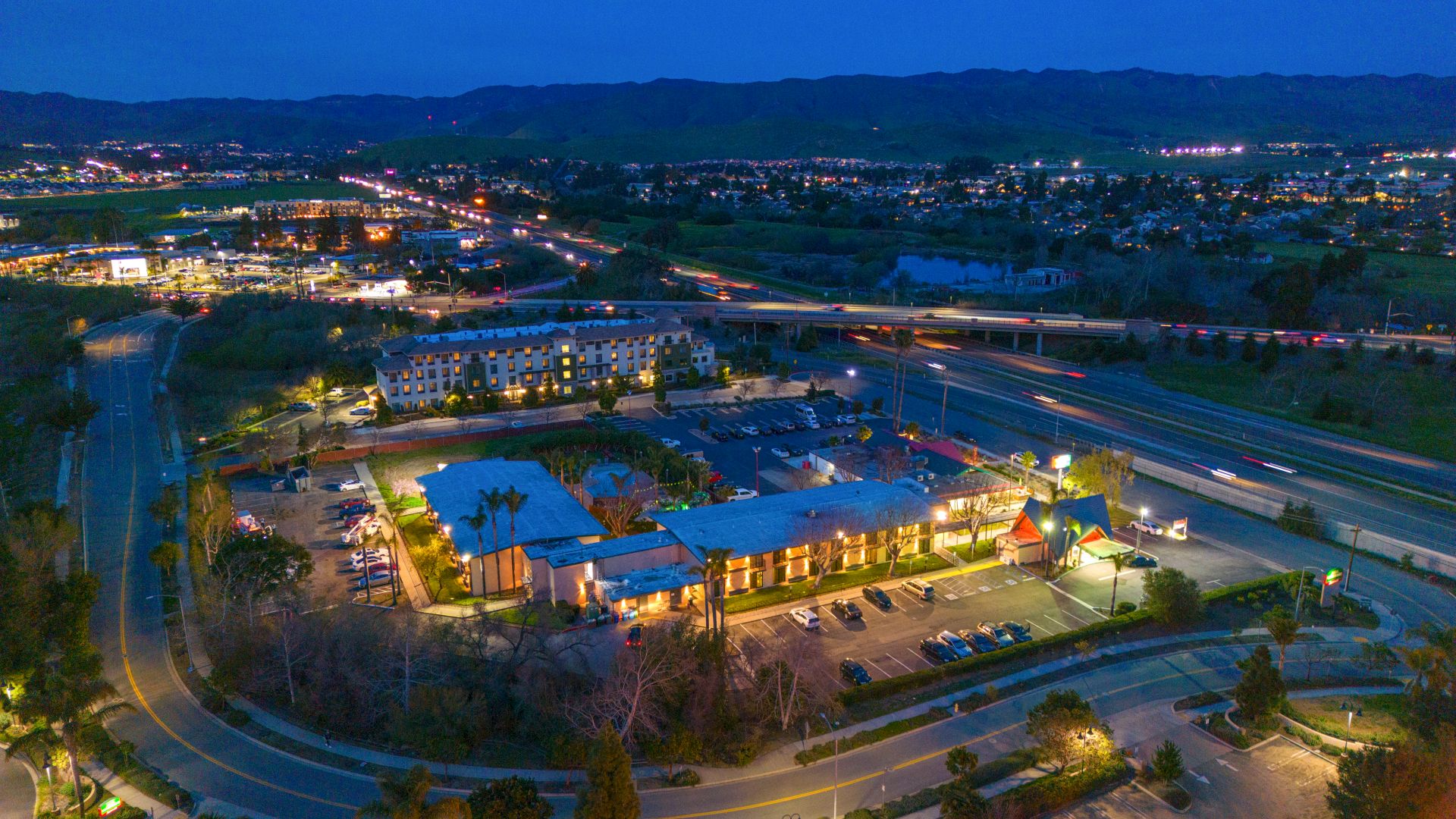 An aerial view of a small city at dusk featuring roads, illuminated buildings, parking lots, and a backdrop of mountains.