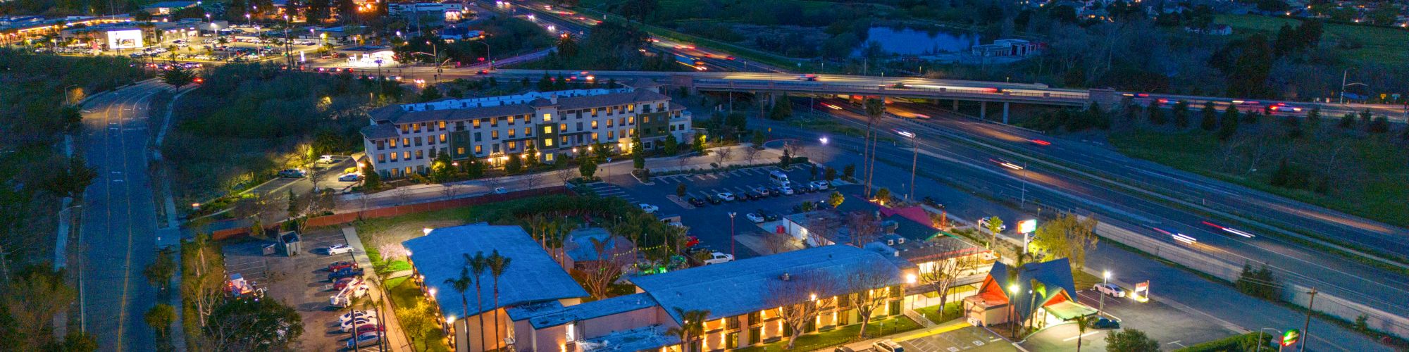 An aerial view of a small city at dusk featuring roads, illuminated buildings, parking lots, and a backdrop of mountains.