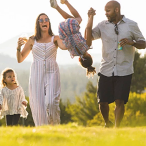 A family of four is enjoying an outdoor activity, with two parents swinging a child playfully between them and another child walking beside them.