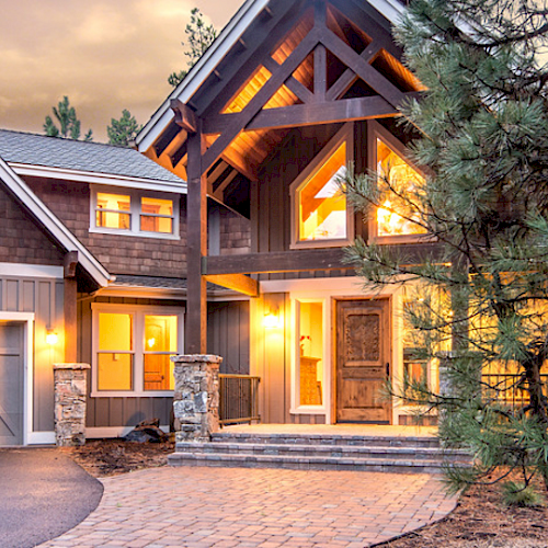 The image shows a large, well-lit wooden house with multiple peaked roofs, large windows, and an attached three-car garage, surrounded by trees.