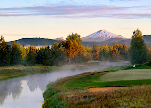 A serene landscape with a golf course near a river, surrounded by trees and mountains in the background, under a clear sky with soft sunlight ending the sentence.