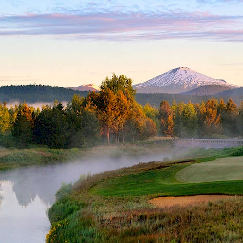 A serene landscape with a golf course near a river, surrounded by trees and mountains in the background, under a clear sky with soft sunlight ending the sentence.
