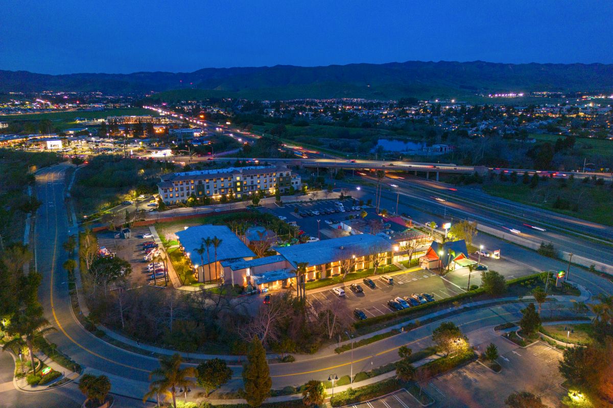 Aerial view of a hotel building with parking lot and surrounding landscape at dusk.