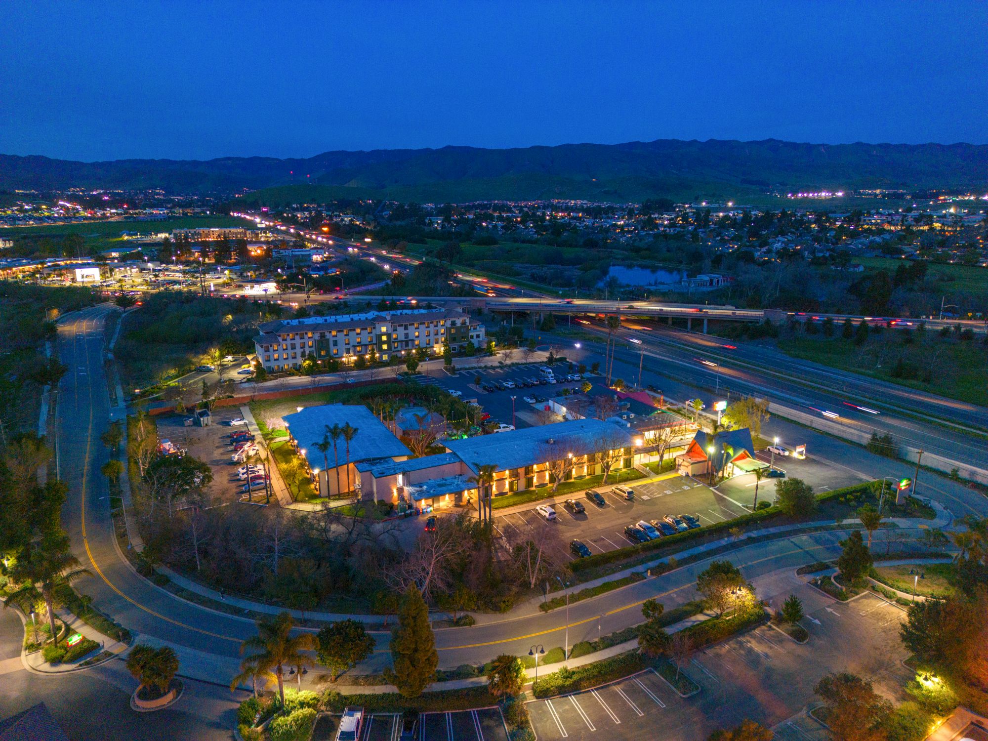 An aerial view at dusk of a suburban area with illuminated buildings, roads, and surrounding greenery, set against a backdrop of hills and highways.