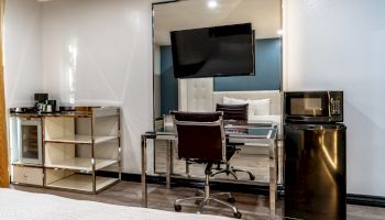 The image shows a modern hotel room with a desk, chair, wall-mounted TV, microwave, mini-fridge, and open shelving.