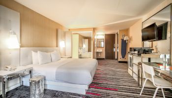 A modern hotel room with a king-sized bed, wall-mounted TV, desk, chair, and stylish decor in neutral tones with red accents.