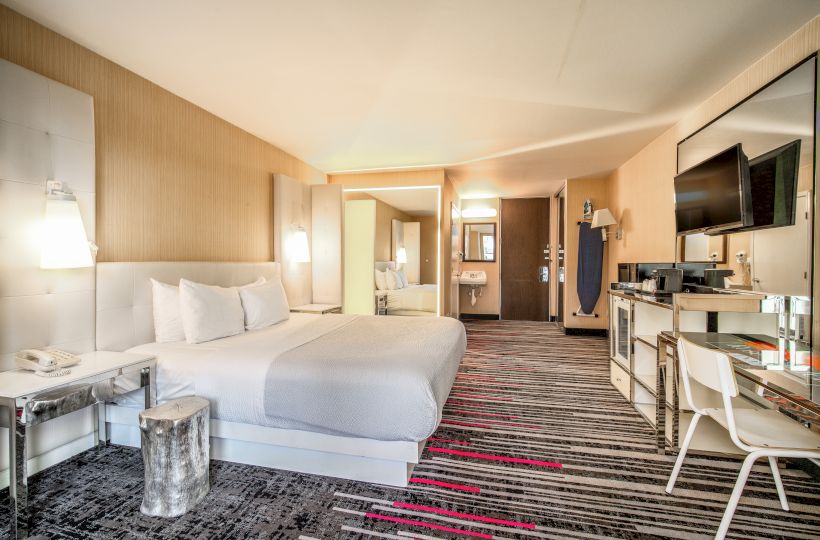 A modern hotel room with a king-sized bed, wall-mounted TV, desk, chair, and stylish decor in neutral tones with red accents.